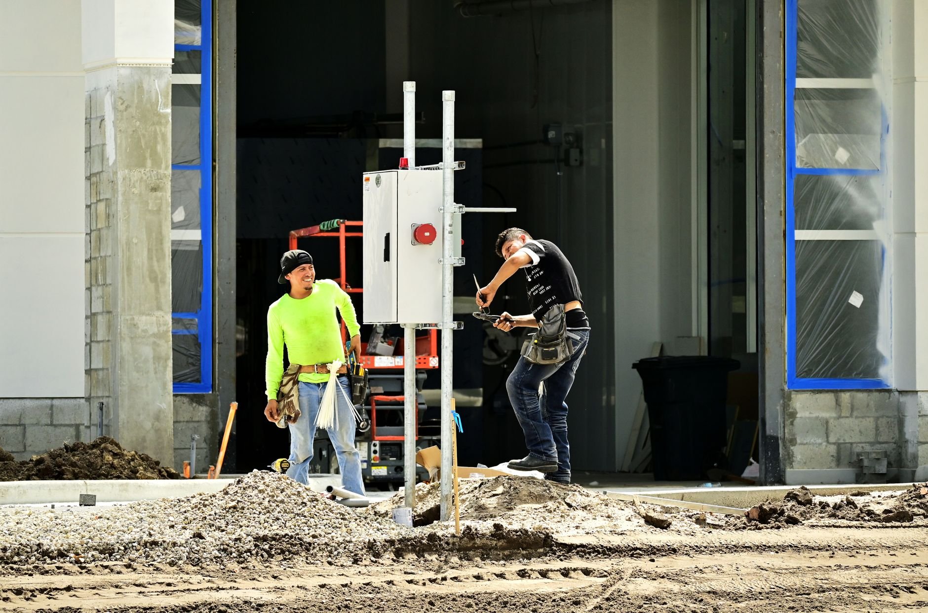 Two construction workers install an electrical panel outside a building site, wearing safety gear.